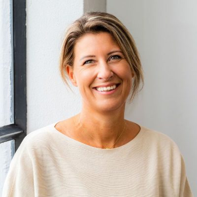 Vrouw met lichte huid en blonde haren die glimlacht bij natuurlijk daglicht, staand naast een grote raam, binnen, casual kleding, vrolijke uitstraling.