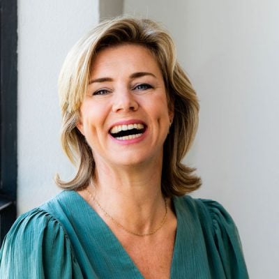 Licht, portret van lachende vrouw met korte blonde haar, natuurlijke uitstraling, kleurrijk blouse, binnenomgeving, fris en vrolijk gezicht.