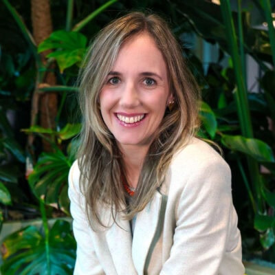 Stralende vrouw met lang, golvend blond haar en groene ogen, lachend in een groene, botanische achtergrond met grote bladeren, gekleed in een witte blazer voor professionele uitstraling.