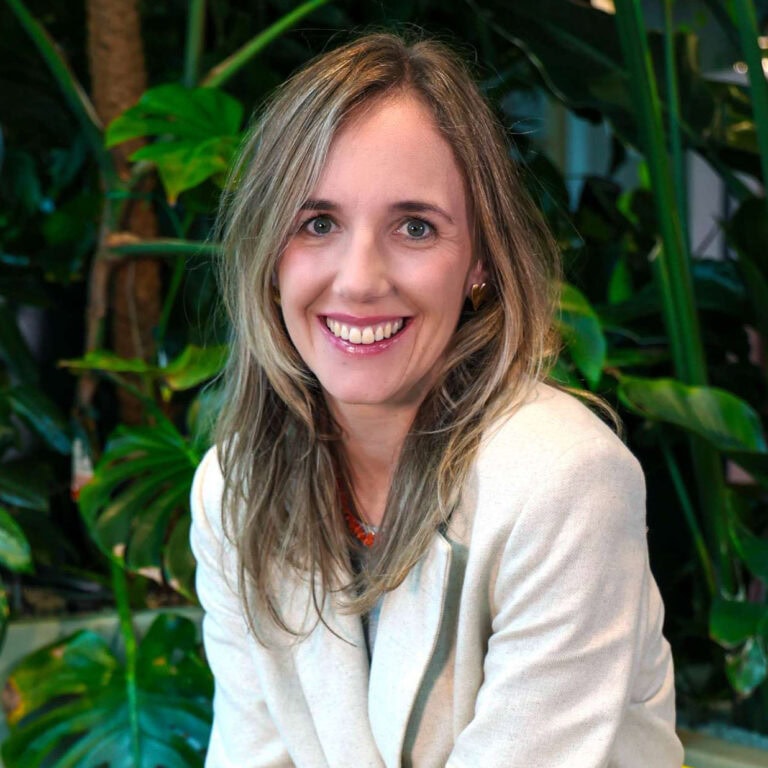 Stralende vrouw met lang, golvend blond haar en groene ogen, lachend in een groene, botanische achtergrond met grote bladeren, gekleed in een witte blazer voor professionele uitstraling.