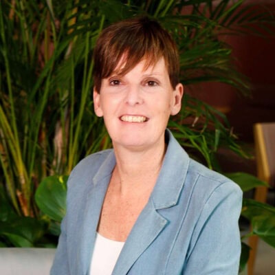 Geconcentreerde vrouw met kort bruin haar in een lichte blazer en witte blouse, zittend voor grote groene kamerplanten, professioneel portret voor inspirerende zakelijke omgeving.