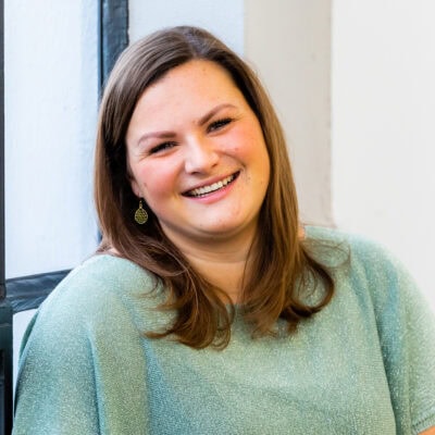 Vrolijke jonge vrouw met bruine haar en oorbellen, glimlachend voor een lichte achtergrond, straalt warmte en authenticiteit uit, perfect voor gebruik op persoonlijke of professionele websites.