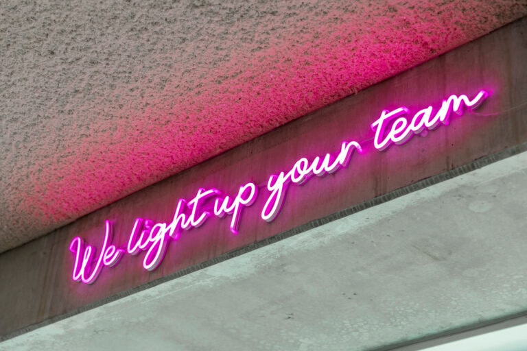 Kleurrijke neonlichttekst op muur met de woorden "We light up your team" in pink.