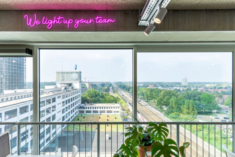 Kleurrijke neonletters die ‘We light up your team’ spellen, hangen boven grote ramen met uitzicht op een stad met groene bomen, pyramide-achtige gebouwen en een spoorweg, in een modern kantoor.
