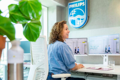 Vergroening door een grote groene plant in een kantoor met een vrouw die aan een computer werkt, met het LICHT logo op de muur.