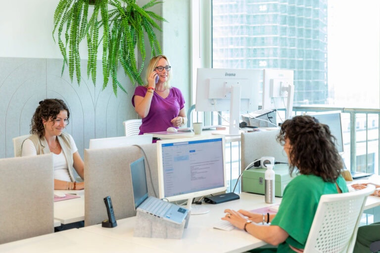 Breda-Leidschendam, Nederland, een moderne kantoorruimte met drie vrouwen die samenwerken en telefoneren, met grote ramen en groene planten, onderstrepen een frisse en productieve werkomgeving.