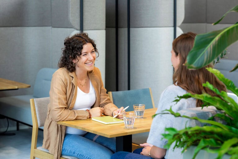 Een vrouw met curly haar in een beige jas spreekt met een andere vrouw met bruin haar in een modern kantoor. Ze lachen en maken oogcontact tijdens een gesprek; er staan twee glazen water, een notitieboekje en pennen op de tafel.