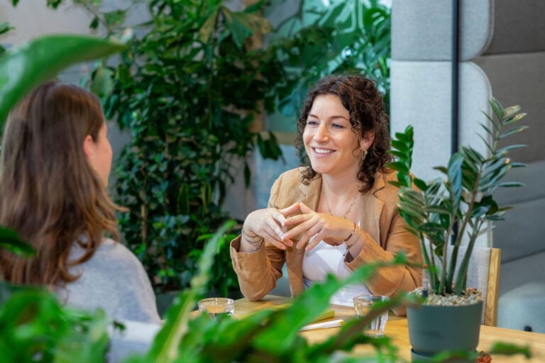 Gezichten van twee vrouwen die een gesprek voeren in een groene, natuurlijke sfeer, omringd door planten.