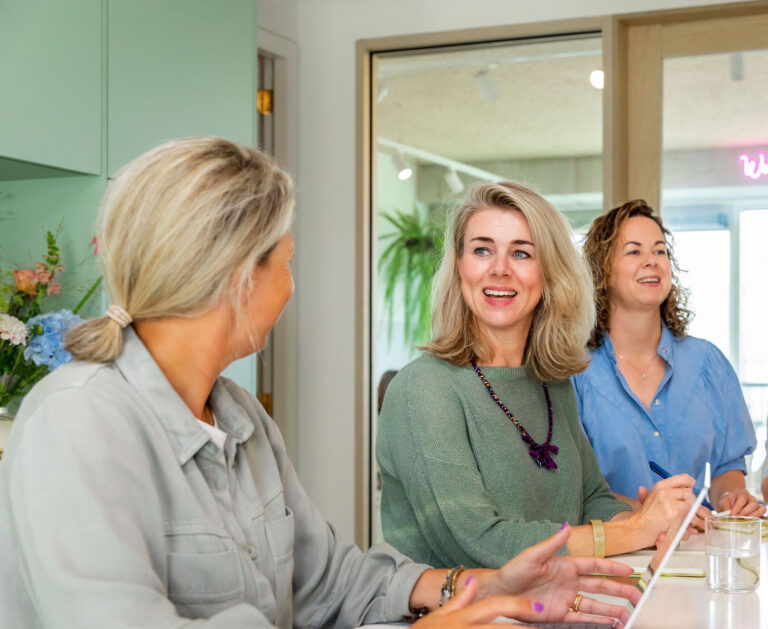 Gezellige zakelijke bijeenkomst met vrouwen in een moderne, lichte vergaderruimte, gericht op samenwerking en communicatie, met bloemen en planten voor een frisse sfeer.