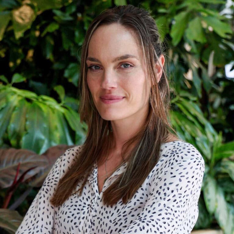 Sfeervolle portretfoto van een vrouw met lang bruin haar en lichte make-up, zittend in een groene, botanische omgeving, benadrukt natuurlijke schoonheid en rust.
