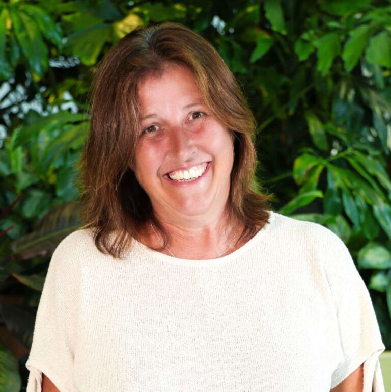 Vrolijke vrouw met brede glimlach en bruin haar, poserend in een groene, geplante omgeving, straalt warmte en vriendelijkheid uit.
