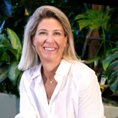 Gezichtsportret van een lachende vrouw met blonde haar, gekleed in een witte blouse, op de achtergrond groen tropisch planten, moderne en frisse uitstraling, geschikt voor gezondheids- en welzijnscontent.