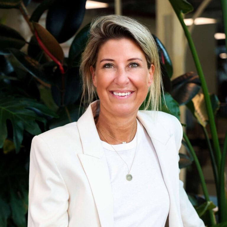 Gezichtsportret van een lachende vrouw in een witte blazer, met groene planten op de achtergrond, geschikt voor bedrijfs- of persoonlijke coaching SEO.