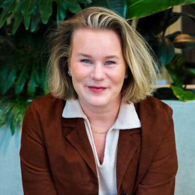 Stralende vrouw met blond haar en blauwe ogen, zittend op een lichte bank met grote groene planten op de achtergrond, in een natuurlijke en frisse kantooromgeving.