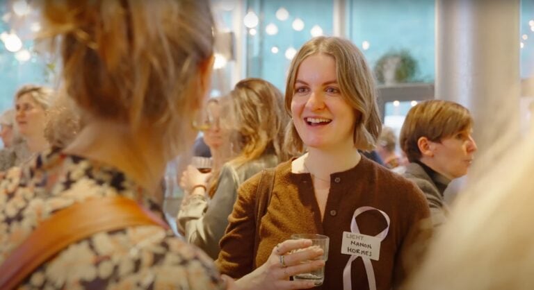 Lichtt, netwerkevenement, sociale interactie, vrouw met naamlabel Manon Hornes, glimlach, professionele bijeenkomst, trainer, communicatie, inspireren, professionele groei, vrouw met glas drinken, vriendelijke sfeer, netwerkborrel, zakelijke bijeenkomst.
