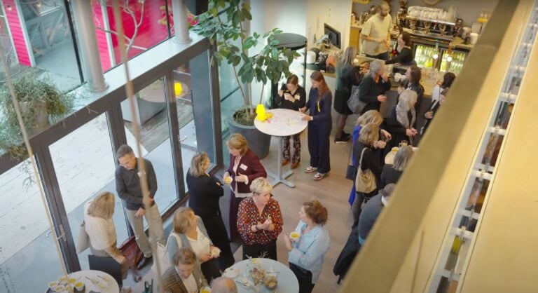 Zicht op een gezellige binnenruimte van LICHT met mensen die genieten van een netwerkbijeenkomst of evenement, gekleed in zakelijke en casual kleding, met een bar en grote ramen die natuurlijk licht binnenlaten.