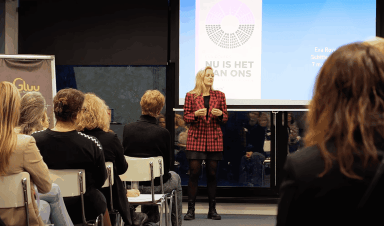 Lichtkunst installatie met publiek en spreker tijdens lezing over inclusiviteit en feminisme, in een moderne setting met grote schermen en branding, voor een inspirerend evenement.