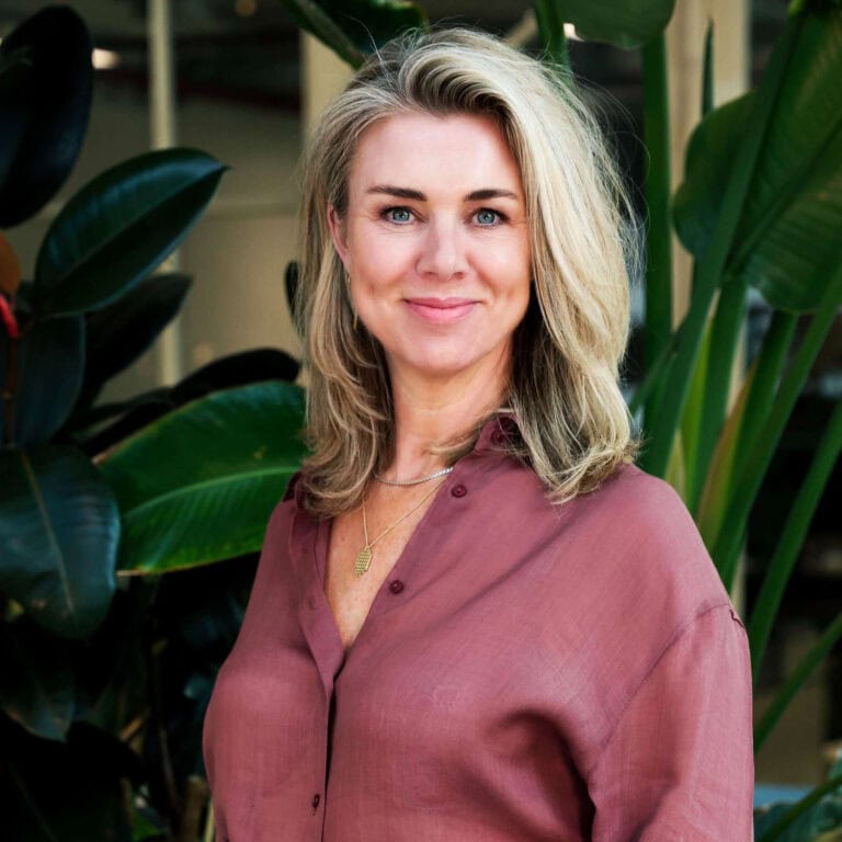 Vrouw met blonde haar en blauwe ogen die glimlacht, gekleed in een roze blouse, omgeven door grote groene bladeren, professioneel portret in een natuurlijke setting, sfeer van rust en elegantie, geschikt voor zakelijke en lifestyle doeleinden.