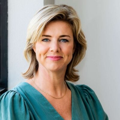 Stralende blonde vrouw met blauwe ogen in een teal blouse, lachend en zelfverzekerd, close-up portret voor een witte muur met natuurlijke verlichting.