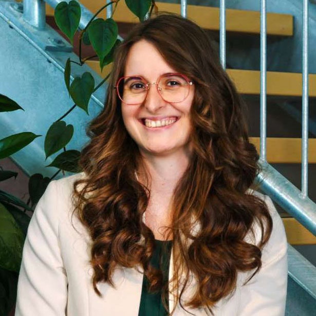 Vrouw met lange krullen, bril en witte blazer lachend, indoors, groene planten en trap op de achtergrond, professionele uitstraling, zakelijke fotografie, modern en vriendelijk, zakelijke vrouw, portret, kleurrijk, menselijke professional, informatief, eigentijds, motivatie, succes, communicatie, leiderschap, werken, bedrijfskantoor, technologie.