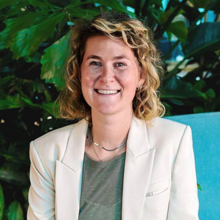 Een vrolijke vrouw met korte, gekrulde blond haar die lacht in een groene botanische setting, draagt een witte blazer en sieraden, stralend en professioneel.