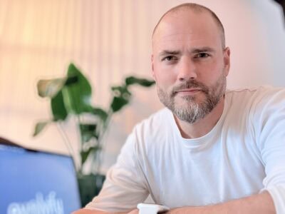 Zachtaardige man met een kort baard en een lichte glimlach, zittend aan een tafel met een laptop en een plant op de achtergrond, in een lichte en rustige interieuromgeving.