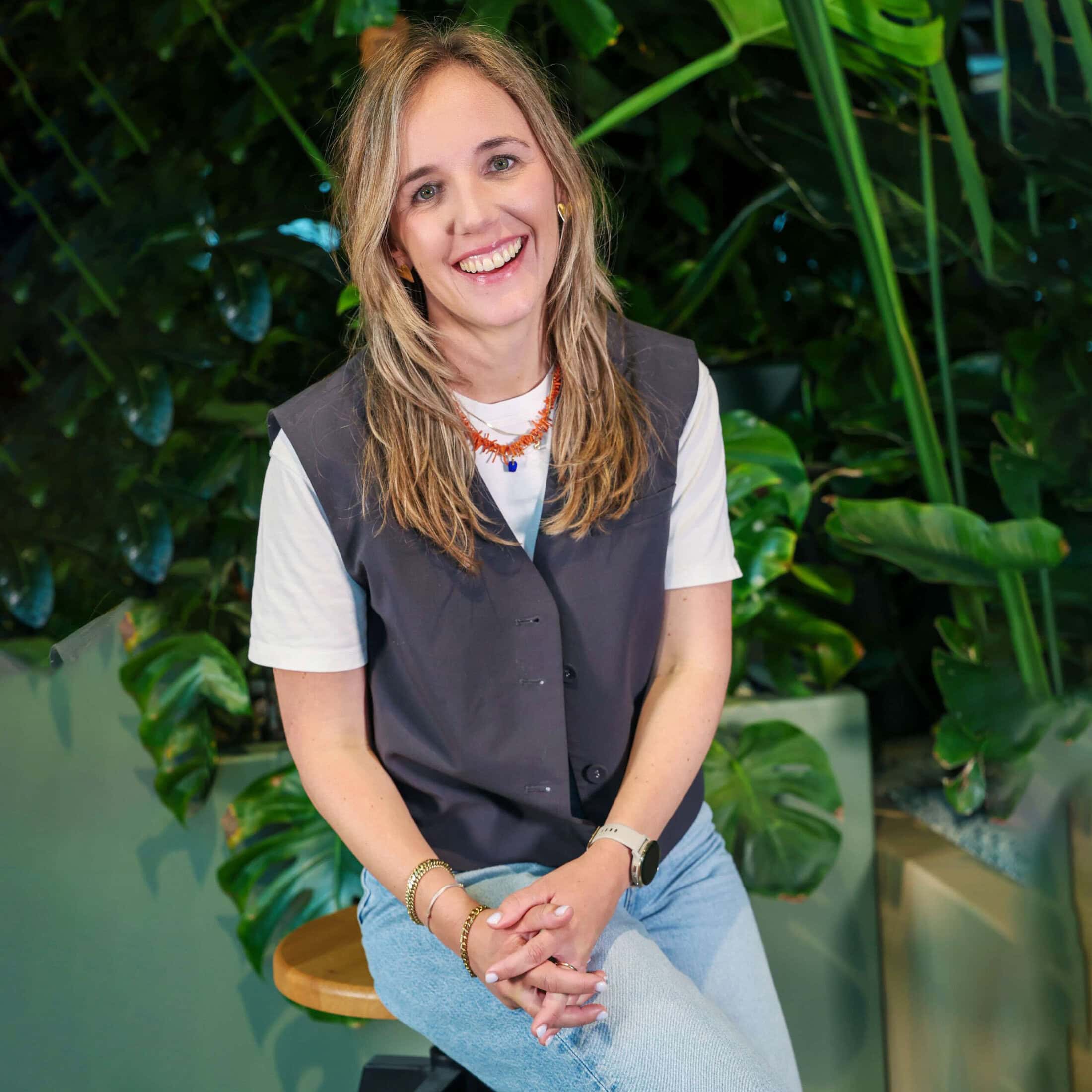 Een enthousiast meisje met lang haar, lachend en zittend op een houten krukje in een groene plantenachtergrond, straalt warmte en positieve energie uit, perfect voor lifestyle en groen beleving.