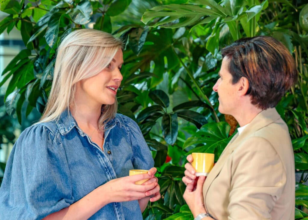 Zachte verlichting en groene achtergrond creëren een warme sfeer tussen twee vrouwen die genieten van een kopje koffie of thee, passend bij het thema LICHT en lichtinval in interieur.