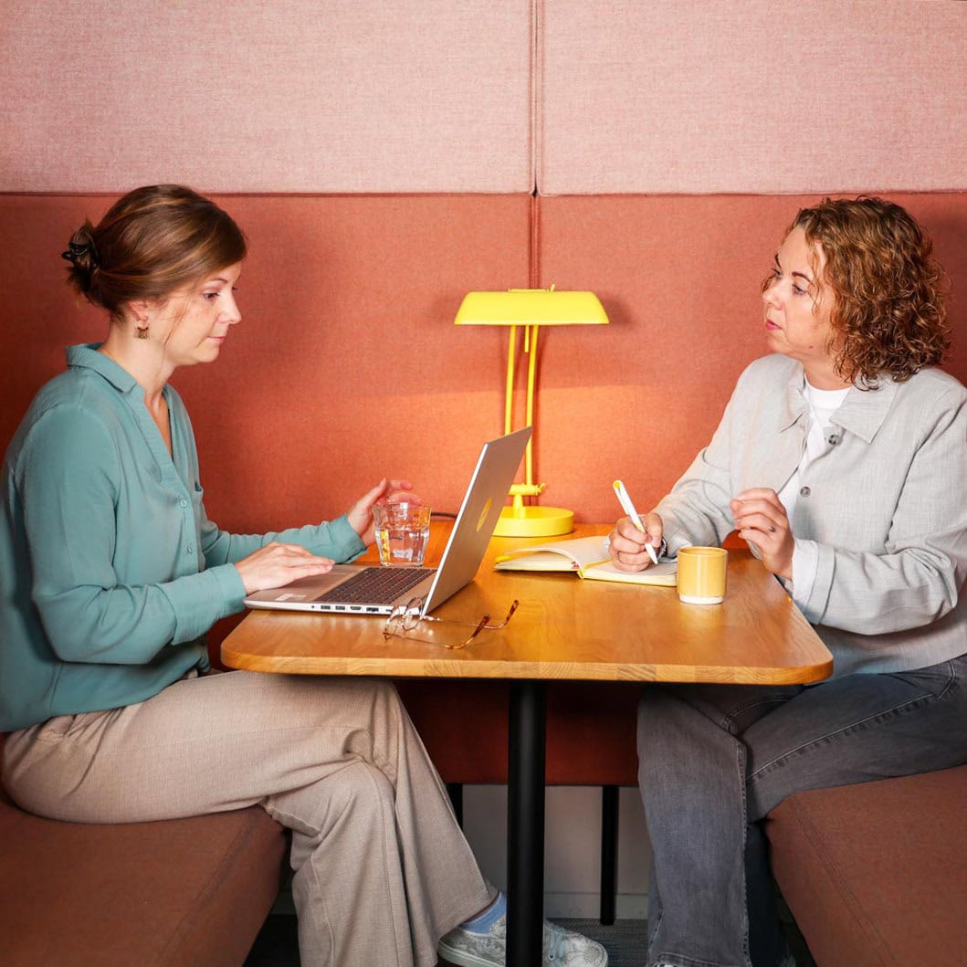 Gezicht aan gezicht gesprek tussen twee vrouwen in een gezellige, lichte kantooromgeving met sfeervol licht en warme kleuren.