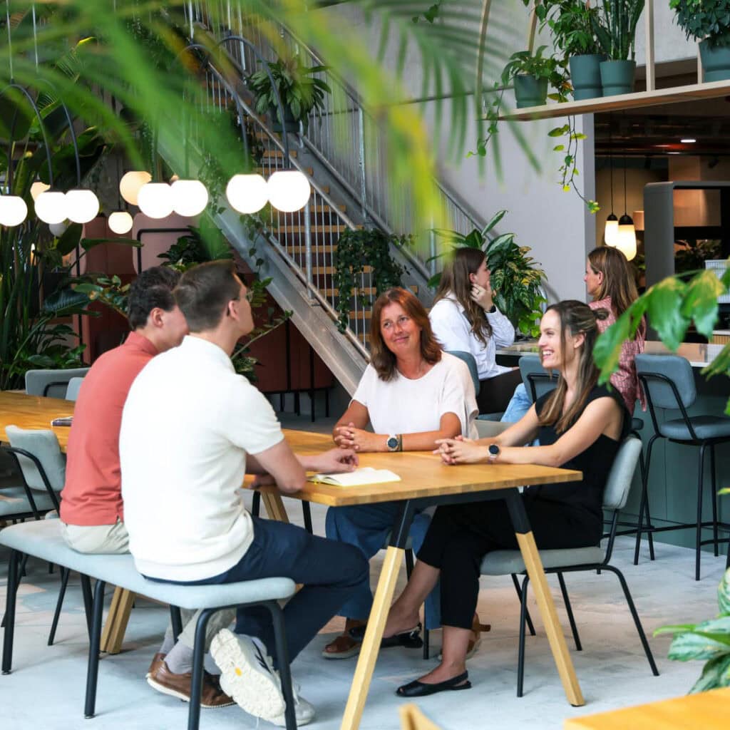 Grote groene planten in een moderne, levendige coworking-ruimte met mensen die in groepsgesprekken zitten en werken.