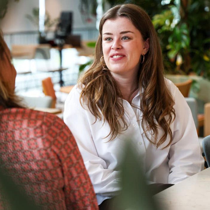 Vrouw in wit overhemd die in gesprek is in een lichte, natuurlijke indoor omgeving met groene planten, symboliseert warmte en menselijke connectie onder het thema Licht.