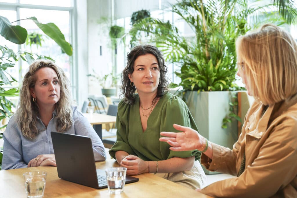 Licht in een moderne kantooromgeving met drie vrouwen die in gesprek zijn, omgeven door groene planten, natuurlijke lichtinval en een inspirerende sfeer voor optimale werkplekken.