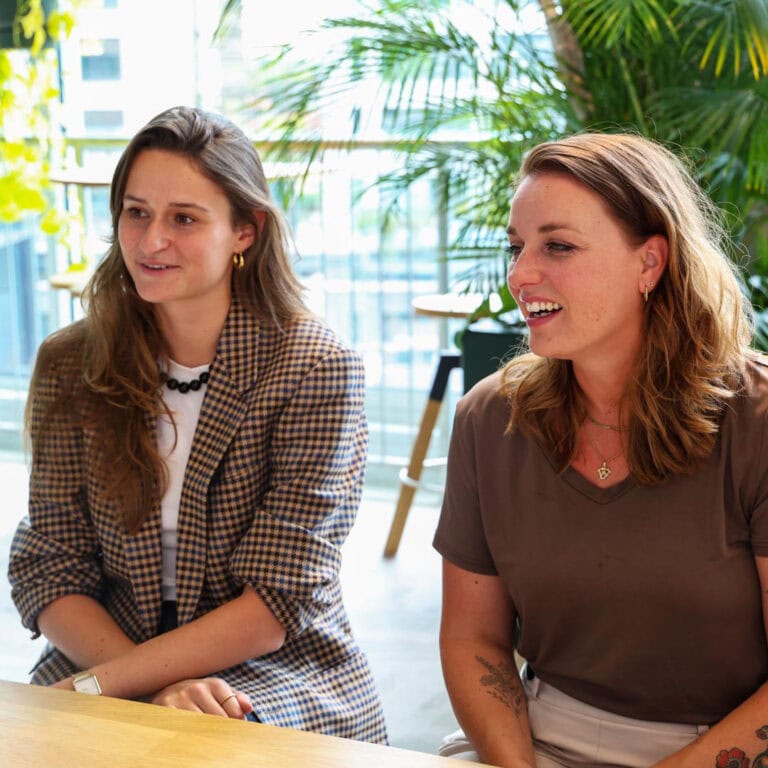 Stralende kantoorverlichting met twee lachende vrouwen in een moderne kantooromgeving, omgeven door groen en natuurlijk licht, ideaal voor innovatieve werkplekken en duurzaam lichtdesign.