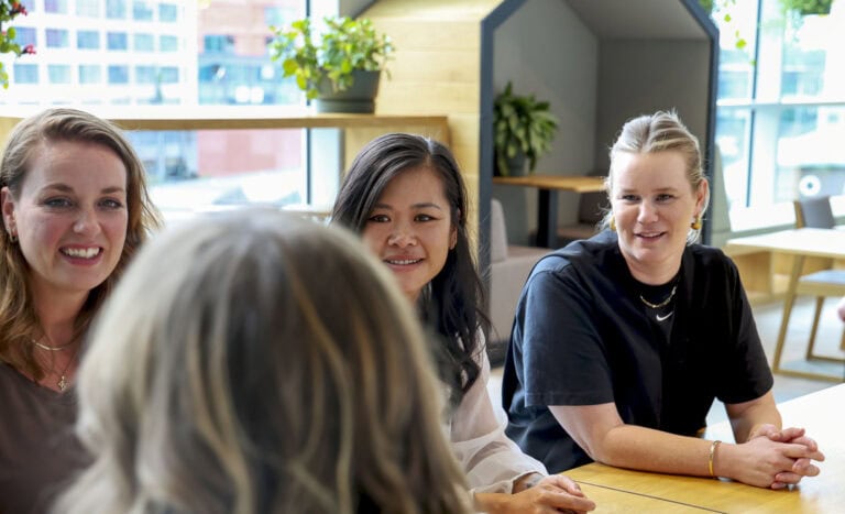 Licht en vergaderingen: vrouwen en man praten en lachen tijdens een creatieve teammeeting in een moderne kantoorruimte met grote ramen, veel groen en natuurlijke lichtinval.