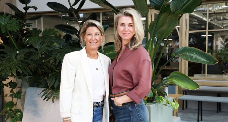 Licht, groen en moderne kantooromgeving met twee vrouwen poseerend tussen grote groene planten voor een inspirerend en natuurlijk werkplekkarakter.