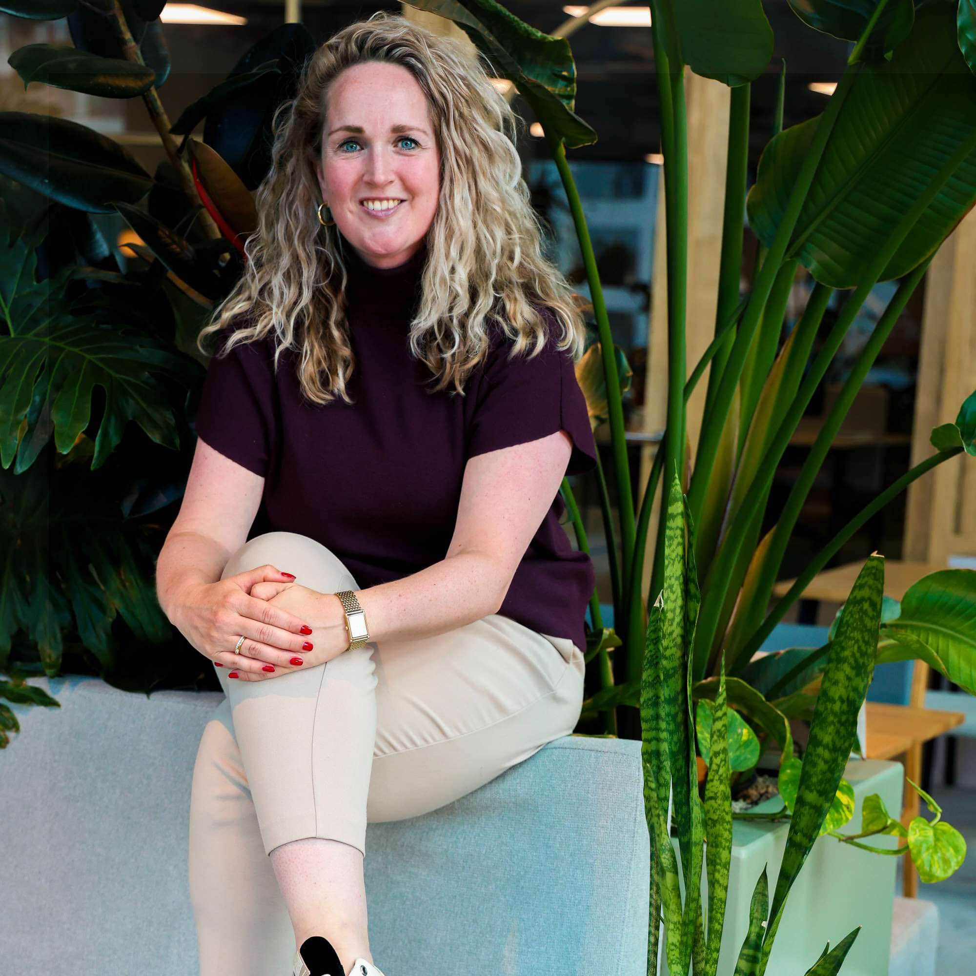 Gezicht van een lachende vrouw met blonde, golvende haren, zittend tussen grote groene planten in een modern interieur, geïnspireerd door licht en natuurlijk groen, passend voor promotie van interieurdecoratie en plantenliefde.