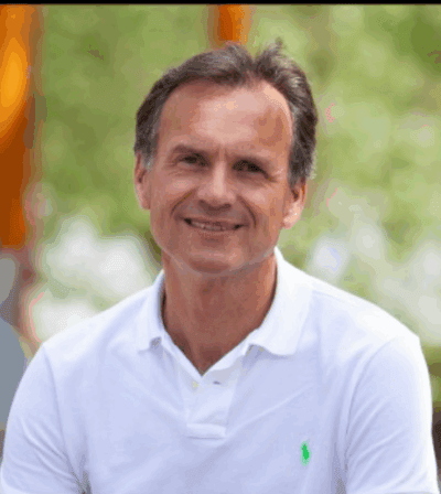 Gezicht van een lachende man met grijs haar, gekleed in een wit polo, buiten in een groene omgeving, stralend zonlicht en een ontspannen sfeer.