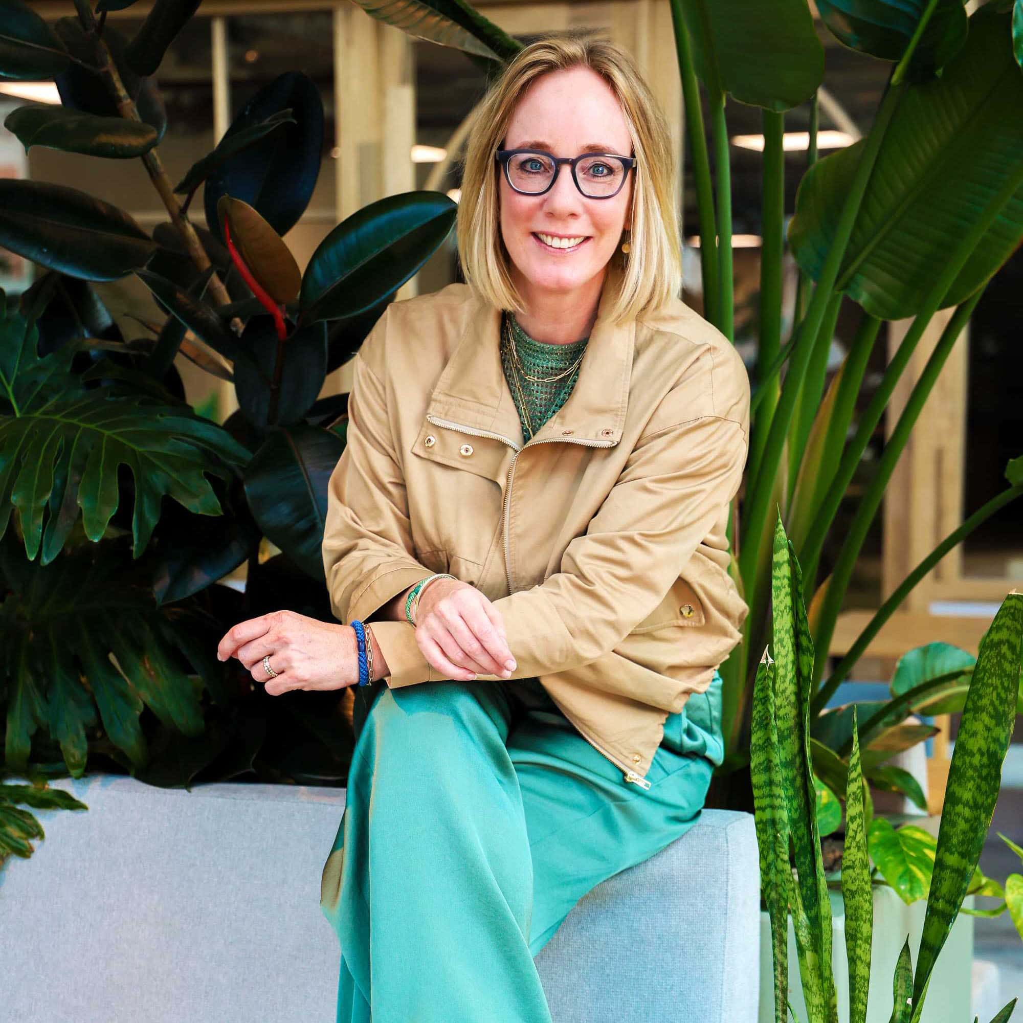 Zonnige vrouw met lichtblonde bob en leesbril zittend tussen grote groene planten in een kantoor of sorteercentrum.