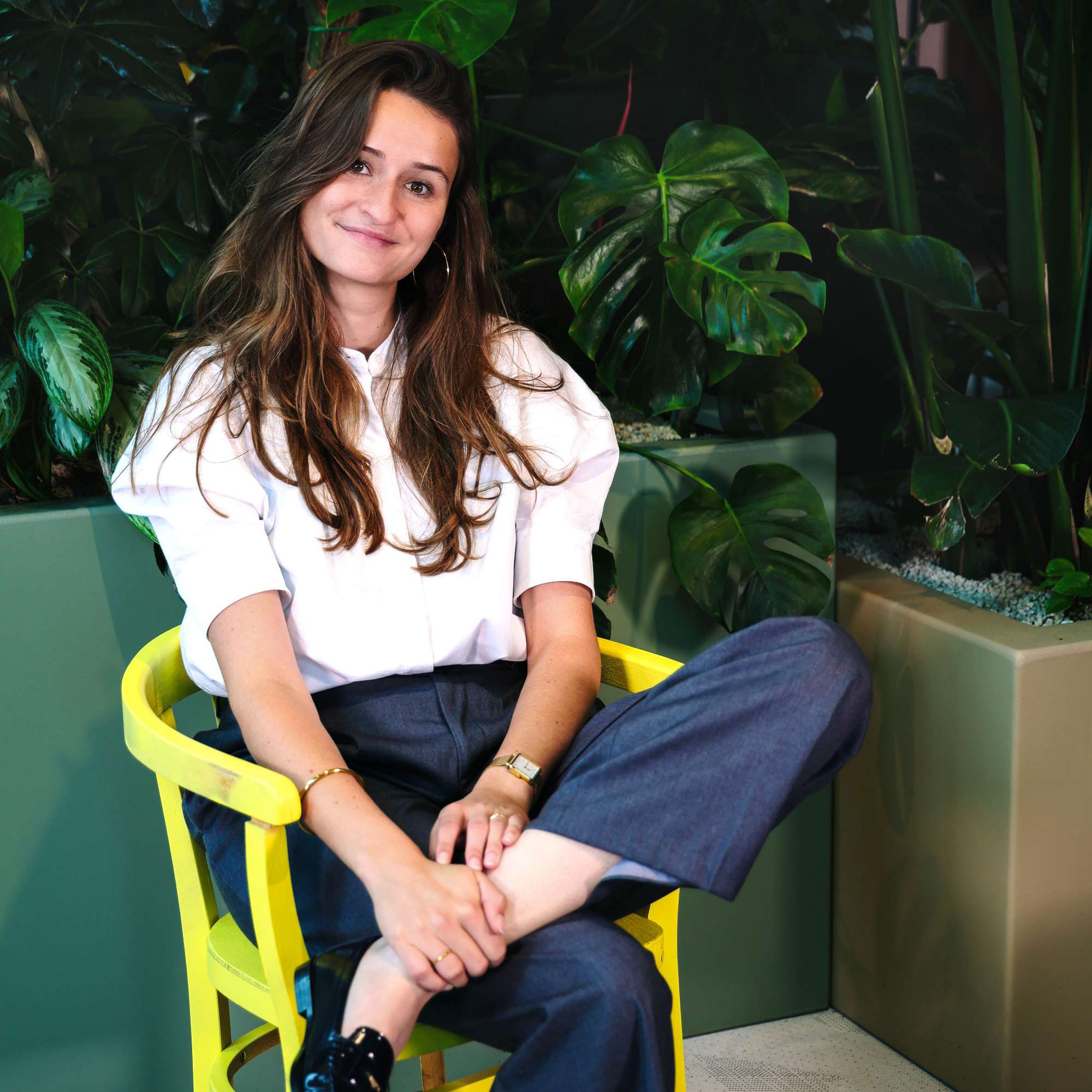 Gezellige vrouw zittend op groene achtergrond met grote monstera bladeren, casual outfit, glimlach, ontspannen sfeer, indoor planten, modern interieur.